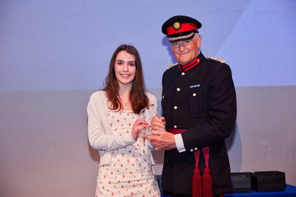 A man in uniform on the right holds an award while Ellie Bealing, stands on the left