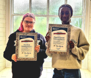 Kira and Abi holding their certificates for their work sitting on interview panels