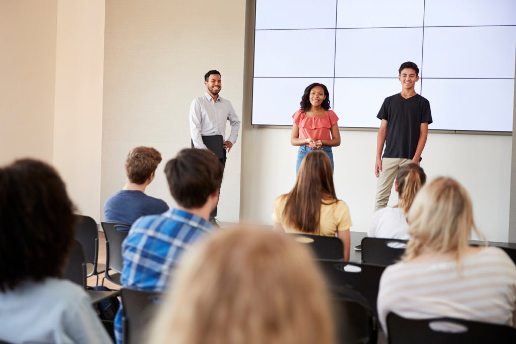 Two young people at the front of a class of students speaking publicly.