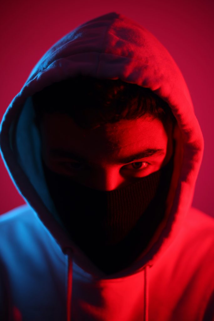 A man in a hoodie with a mask covering his face, partially in shadow and partially with a red light on him.
