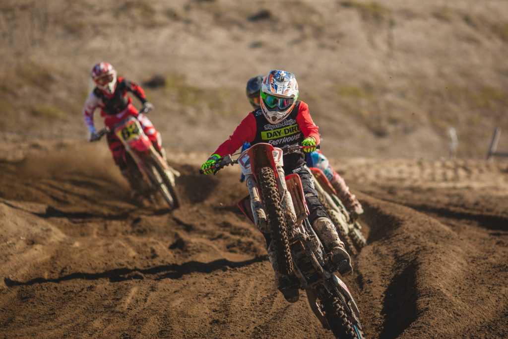A photo of two people on riding motocross bikes towards us on a dusty track and turning to the left.