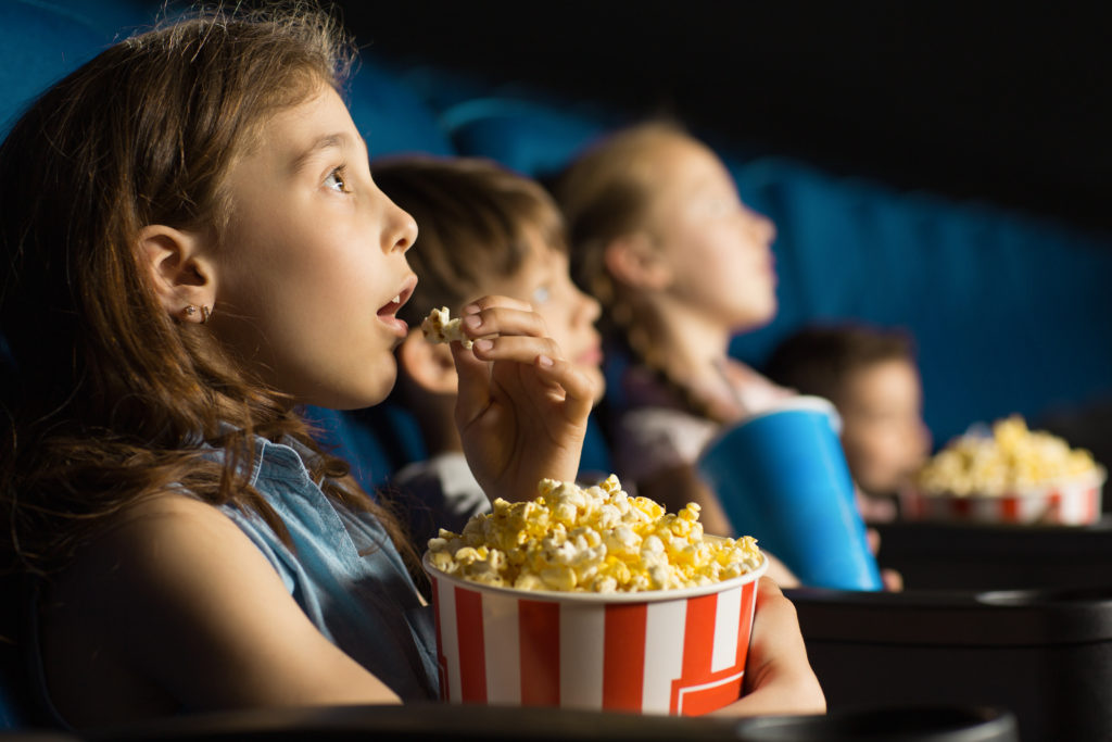 A side view of young people sat in a cinema with awe on their faces and enjoying popcorn.