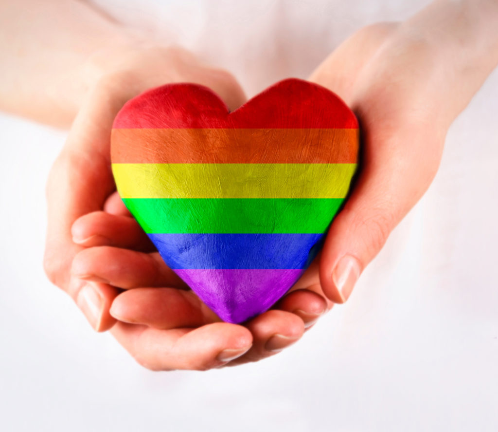 A pair of hands holding a heart painted in the red, orange, yellow, green, blue and purple LGBTQ+ flag colours.
